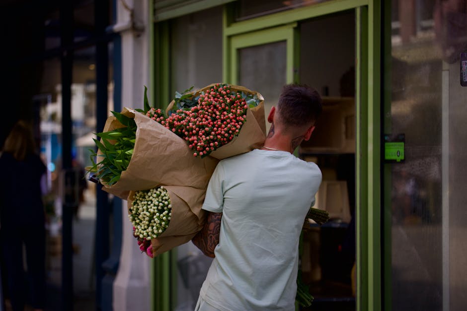 A person with short dark hair and a light-colored T-shirt is carrying a large bouquet of flowers wrapped in brown paper, positioned over their shoulder. The bouquet features pink and white small bloom