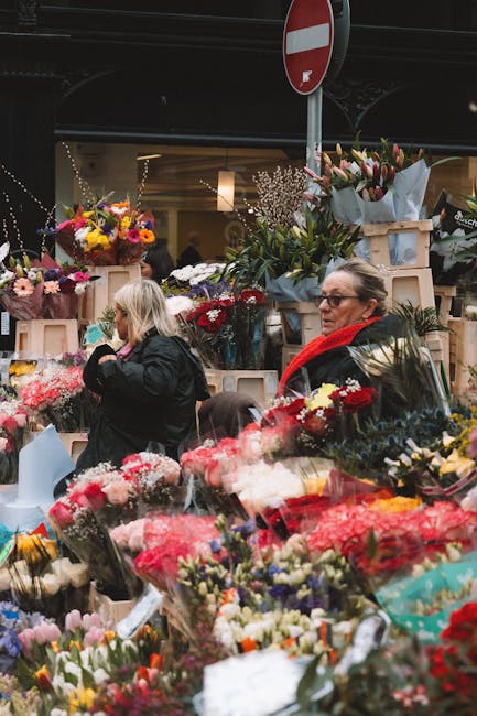 A bustling floral market display featuring a variety of fresh flowers including pink, red, yellow, and white blooms arranged in bouquets and packaging, with some flowers wrapped in colorful paper and 