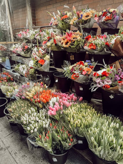 An array of fresh flowers displayed for sale at a florist shop, featuring tulips in shades of pink, white, and red, alongside vibrant orange and purple bouquets. The flowers are arranged in black buck
