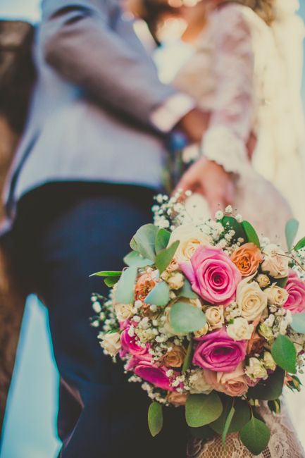 A close-up of a bouquet arrangement held by a bride, featuring pink, peach, and cream roses accented with sprigs of baby's breath and eucalyptus leaves, wrapped in soft beige lace paper with a satin r