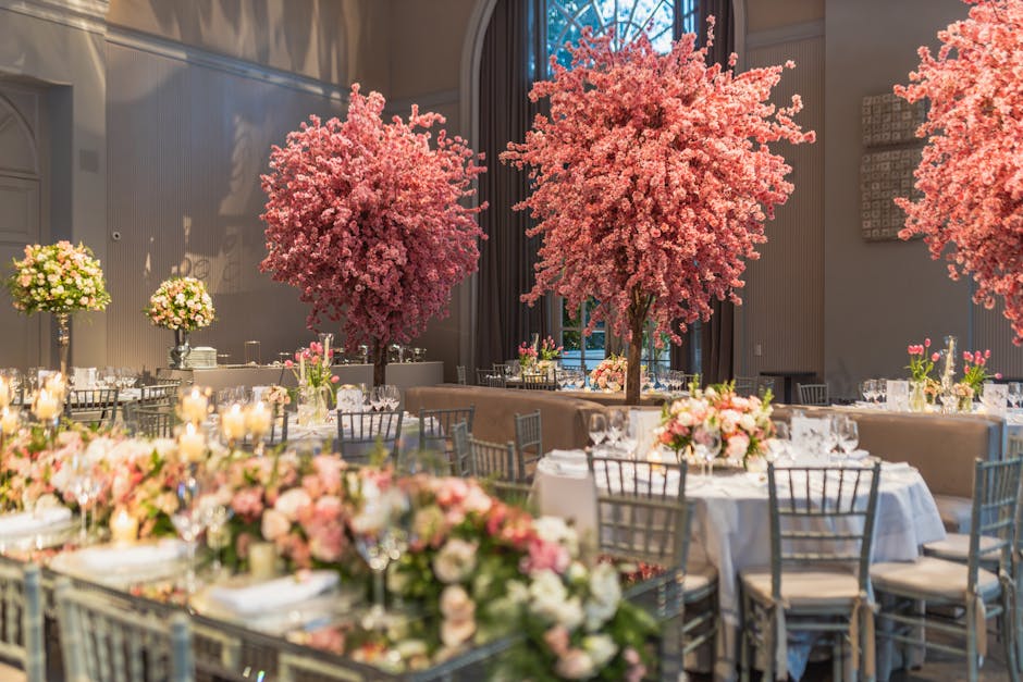 An elegant wedding venue decorated with large, vibrant pink cherry blossom trees and floral centerpieces consisting of pink and white roses, peonies, and greenery arranged in glass vases. The tables a
